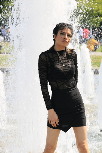 An East Indian model standing in a water fountain getting soaked. She is wearing a black see through top and mini skirt, with a necklace.