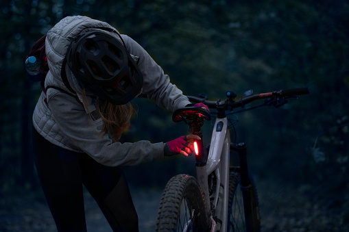 Low-key photo of a cheerful young woman with her enduro mountain bike (eMTB) at night, turning on the rear light.