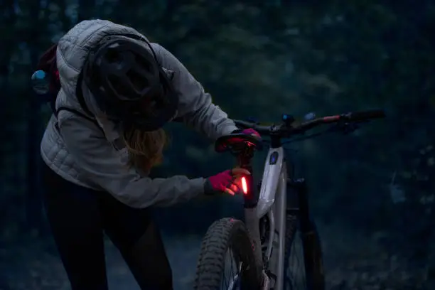 Low-key photo of a cheerful young woman with her enduro mountain bike (eMTB) at night, turning on the rear light.