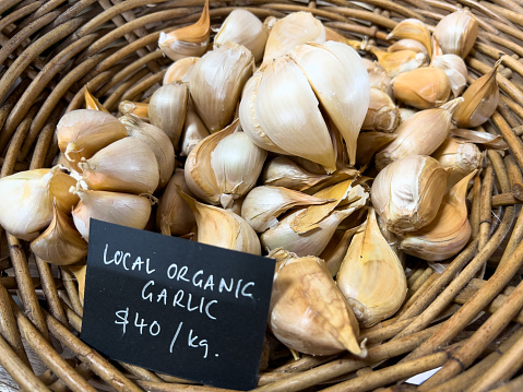Horizontal high angle closeup photo of organic garlic cloves for sale in a woven cane basket with a price tag and description in a delicatessen. Byron Bay, north coast NSW.