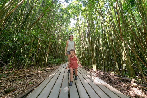 An adorable Eurasian toddler girl laughs while running along a boardwalk trail through a bamboo forest in Hawaii with her mom.