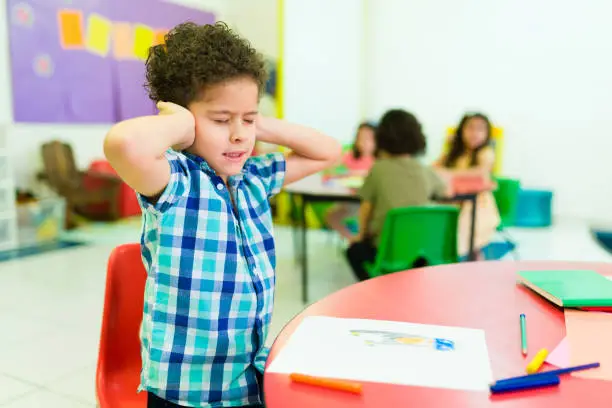Upset autistic little boy covering his ears and feeling distressed and overwhelmed by the loud noises in preschool