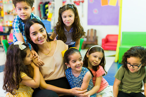 Attractive latin teacher smiling while hugging and playing with her preschool children students in the classroom