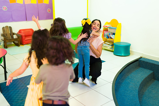 Cheerful preschool teacher welcoming and hugging her happy kindergarten students after coming back to school