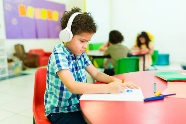 Autistic preschooler with headphones coloring alone in class Sad preschool student boy with autism wearing headphones and feeling isolated from his classmates in kindergarten autism stock pictures, royalty-free photos & images