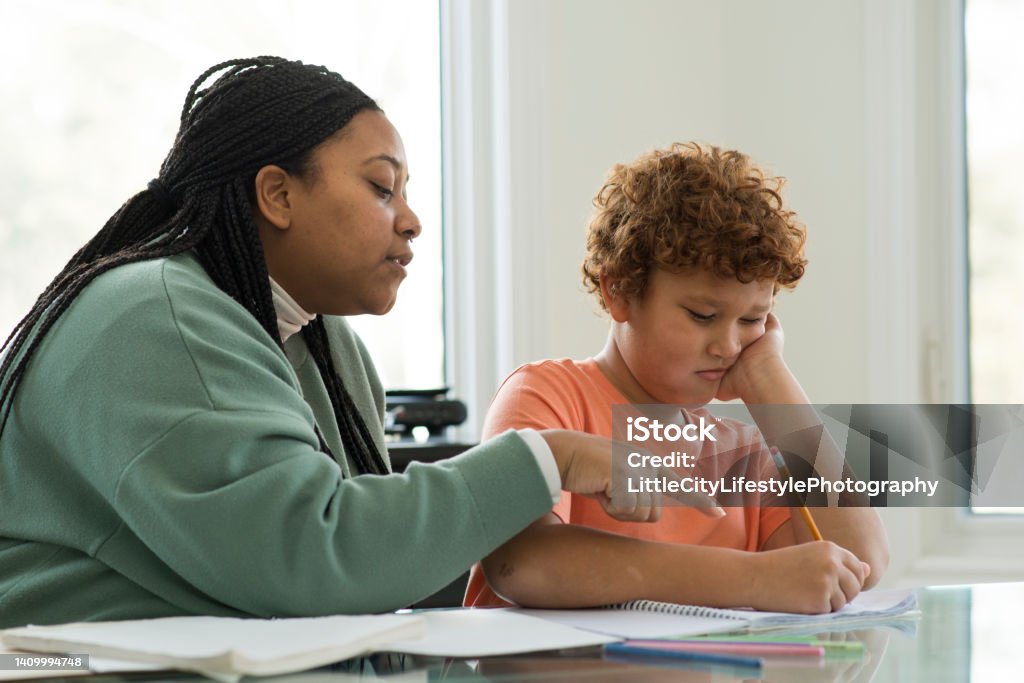 Confusing Homework Young boy is sitting at the kitchen table with his mother while doing homework from school. He has his chin in his hand in frustration with a pencil in the other hand to correct his work in the notebook. His mother is pointing at the words and explaining what needs to be corrected. Struggle Stock Photo