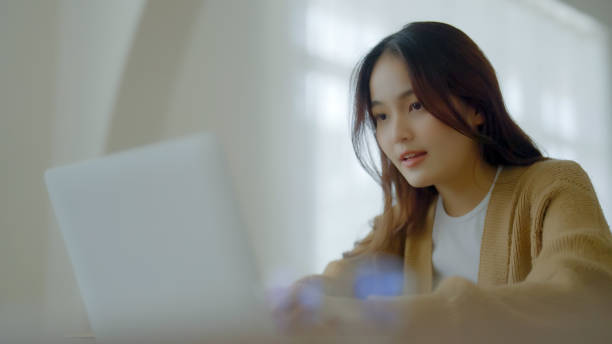 istock Smiling asian young woman working on laptop at home office. Young asian student using computer remote studying, virtual training, e-learning, watching online education webinar at house 1409994794