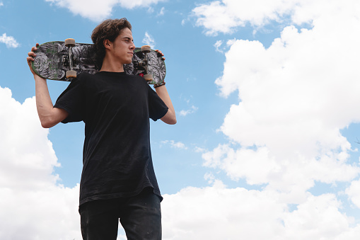 caucasian boy with urban style clutching his skateboard on his shoulders with a blue sky on his shoulders looking into the distance