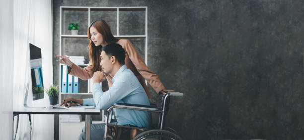 男性は車椅子に座り、同僚、男性と女性の従業員がオフィスで一緒に働き、大きな組織でチームとして働いています。チーム管理の概念は多様です。 - working people physical impairment wheelchair ストックフォトと画像