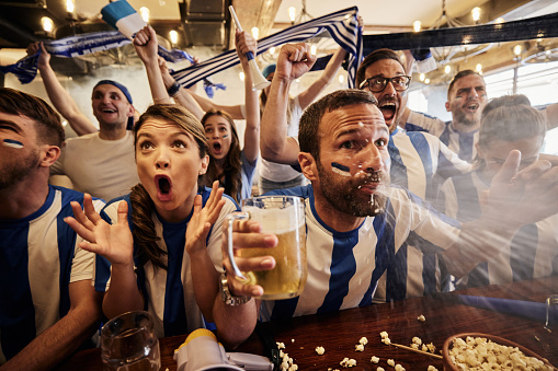 Large group of cheerful sports fans celebrating the success of their team during a game on TV in a bar.