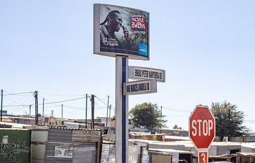 Commercial signs above road signs dedicated to Winnie Madikizela Mandela (late ex-wife of the late Nelson Mandela) and Eneas Peter Nanyemba (Namibian liberation hero 1935-1986) at Hakahana Township near Windhoek in Khomas Region, Namibia. Signs can be seen on the shacks in the background.