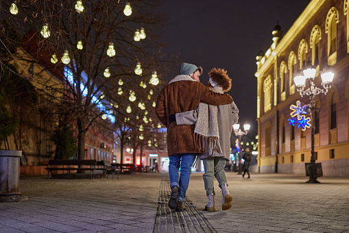 Back view of young happy couple talking while walking embraced during Christmas night on the city street.