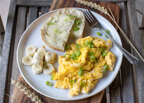 Savory breakfast, old fashioned style with homemade scrambled eggs, sliced white bread with butter and bananas. Served on a plate on wooden background