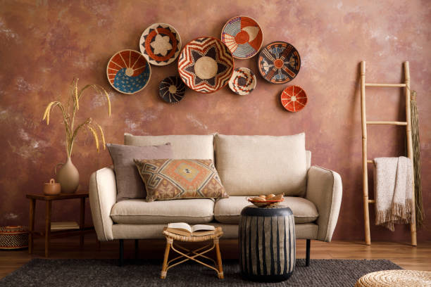 The stylish boho compostion at living room interior with design beige sofa, coffee table, stool wicker baskets and personal accessories. Brown and white pillows and plaid. Cozy apartment. Home decor. el elegante compostaje boho en el interior de la sala de estar con sofá beige de diseño, mesa de centro, cestas de mimbre de taburete y accesorios personales. almohadas marrones y blancas y plaid. acogedor apartamento. decoración del hogar.