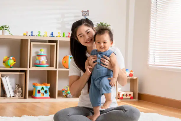 Happy Asian mom kissing her newborn baby smile and spending time for good moment together at warmth place. Adorable little baby boy having fun and cheerful with love from mother.Mother day concept