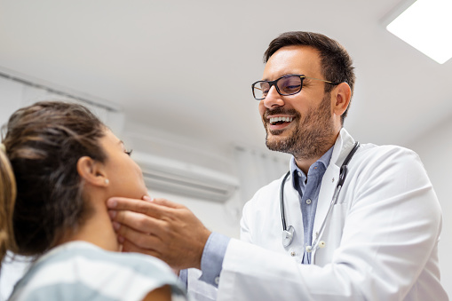 medicine, healthcare and medical exam concept - doctor checking patient's tonsils at hospital. Endocrinologist examining throat of young woman in clinic