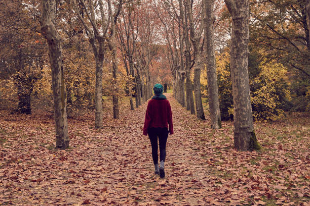 Girl walking backwards along a tree-lined path with brown fallen leaves on the ground in a forest or park in autumn