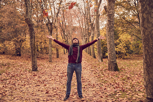 Young white Caucasian man looking upwards throwing leaves from trees in a corridor of trees on brown fallen leaves in the forest with scarf and hat. Autumn colors