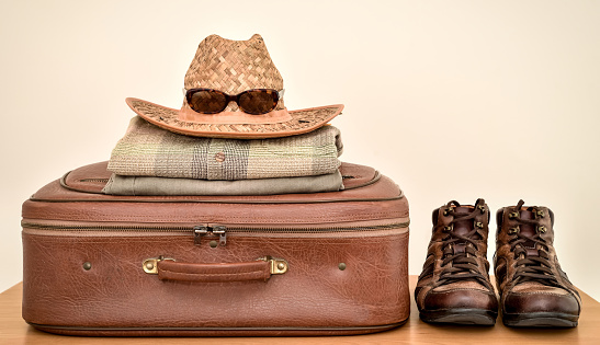 Vintage suitcase, pants, shirt, straw hat, sunglasses and boots on the brown wooden table. Copy space. Travel baggage concept.
