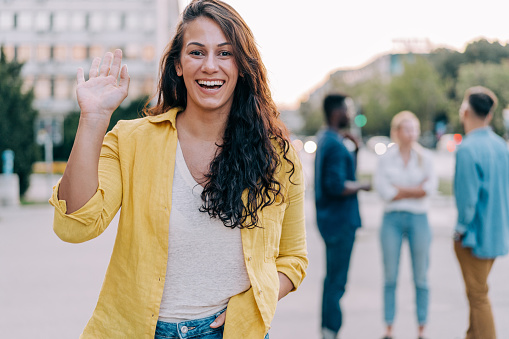 Portrait of beautiful smiling young woman waving with hand and walking on the street.
