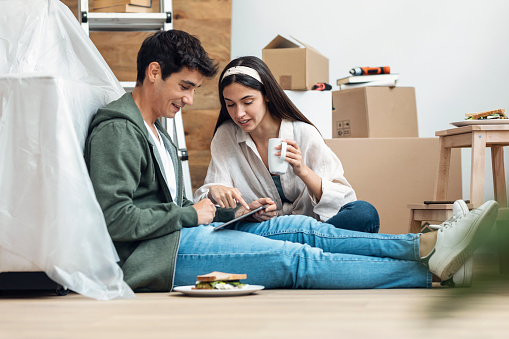 Shot of cute couple using tablet while relaxing from moving in their new house arround cardboard boxes