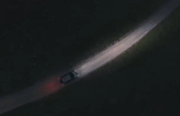 The car moves on a dirt road at night. The road is lit with headlights. Air top view. Theme of road transport