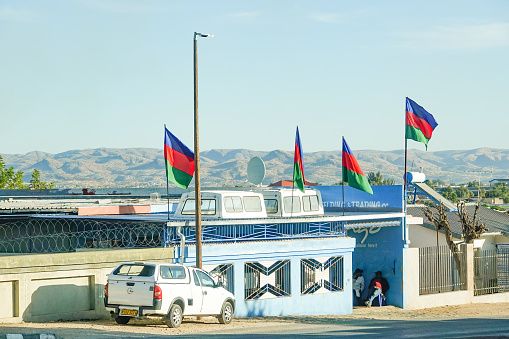 People and car number plates are visible outside a business with the Namibian Flag overhead at Katutura Township near Windhoek in Khomas Region, Namibia