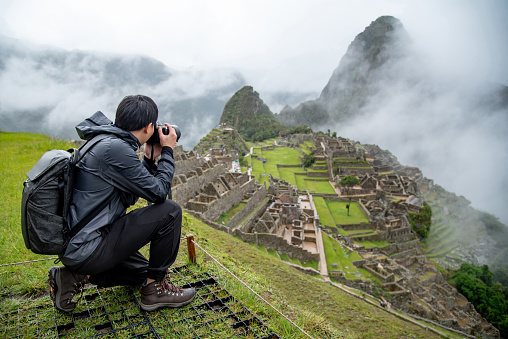 Asian man tourist and photographer taking photo at Machu Picchu, one of seven wonders and famous tourist attraction in Cusco Region of Peru. This majestic place has known as Lost City of the Incas.