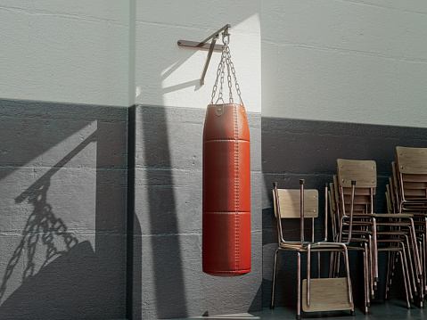 A red leather punching bag mounted on a wall in a room with stacked chairs lit by a window light - 3D render