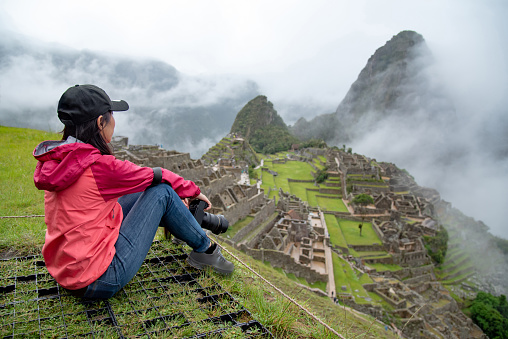 Asian woman tourist looking at Machu Picchu, one of seven wonders and famous tourist attraction in Cusco Region of Peru. This majestic place has known as Lost City of the Incas.