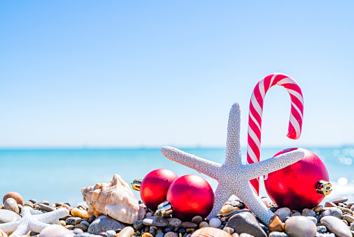 Christmas backgrounds: candy cane, starfish and Christmas ornaments standing on beach pebbles with defocused tropical beach at background. High resolution 42Mp outdoors digital capture taken with SONY A7rII and Zeiss Batis 40mm F2.0 CF lens