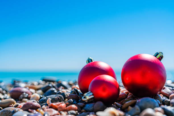 Christmas backgrounds: red Christmas balls on beach pebbles with defocused tropical beach at background. High resolution 42Mp outdoors digital capture taken with SONY A7rII and Zeiss Batis 40mm F2.0 CF lens