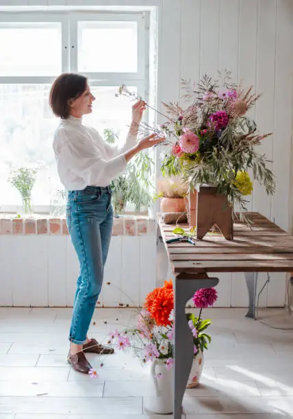 Female professional florist prepares the arrangement of wild flowers. Flower shop. Background concrete gray wall. Concept inspiration, floral, greetings, spring, ornament flowers.