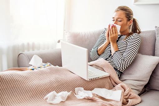 Shot of a woman using a laptop while blowing her nose with a tissue. Woman blowing her nose. Allergic woman blowing nose in tissue sit on sofa at home office study work on laptop