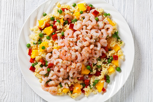 cooked salad shrimps with millet porridge with green peas, mango, red peppers on white plate on white wood  table, horizontal view from above, flat lay, close-up