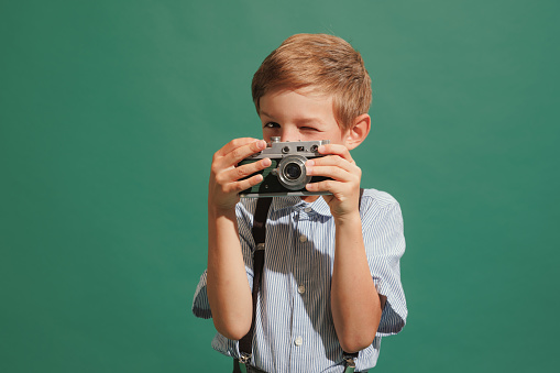 Taking picture with retro phote camera. One preschool boy, kid wearing retro style outfit isolated on dark green background. Concept of child emotions, facial expression, fashion and ad.