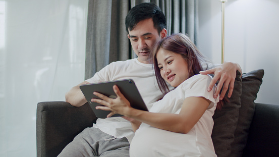 Happy caucasian husband with a pregnant wife looking ultrasound photo of her newborn baby together on a tablet and shopping online in the living room at home.