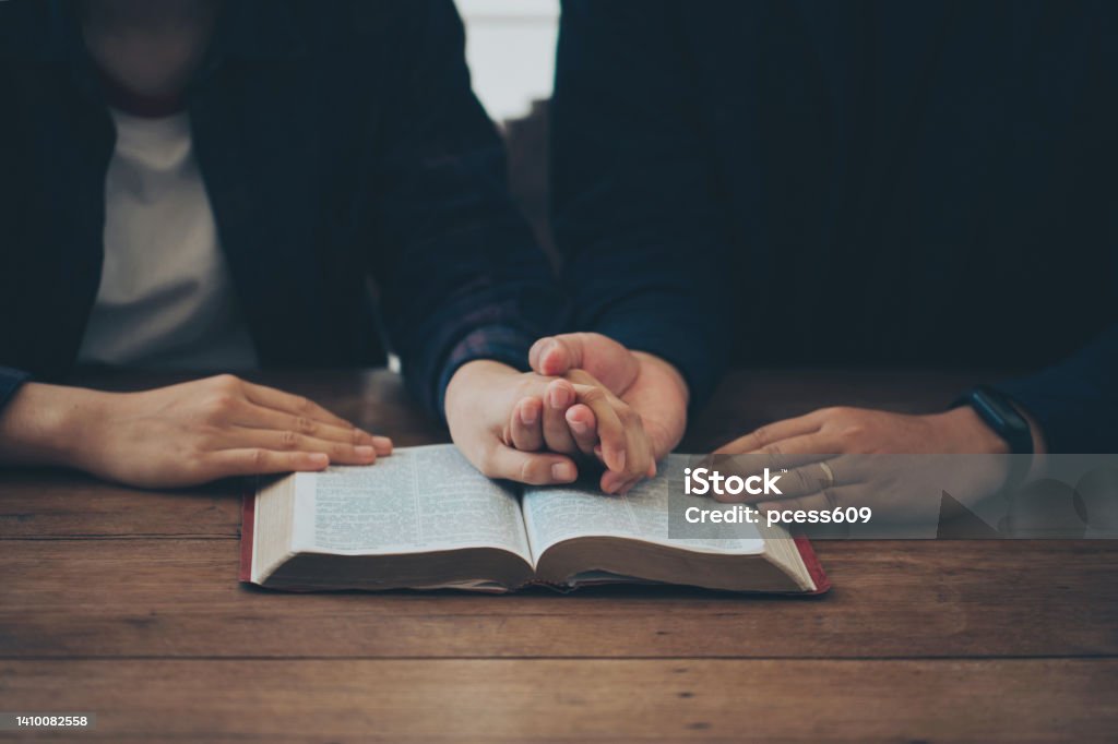 Two Christian couples holding each other's hands praying together over the bible on a wooden table. Begging for forgiveness and believing in goodness. Christian life crisis prayer to god. Couple - Relationship Stock Photo
