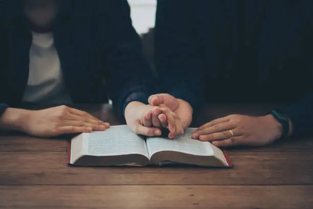 Photo of Two Christian couples holding each other's hands praying together over the bible on a wooden table. Begging for forgiveness and believing in goodness. Christian life crisis prayer to god.