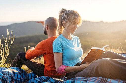 A young couple is relaxing on the mountain. They are sitting on blanket back to back, reading book and enjoying the view.