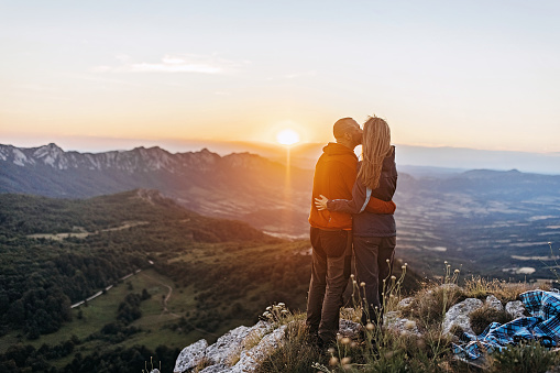 Portrait of a young couple kissing on the cheek, with a beautiful sunrise in front of them.