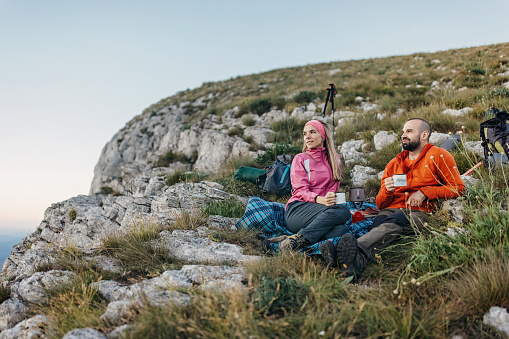 A hiking couple is sitting on top of a mountain, holding tea and looking thoughtfully into the distance.