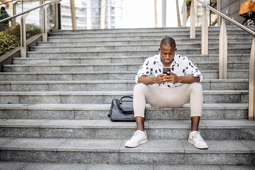 Handsome man using phone while sitting on a public stairs in a city