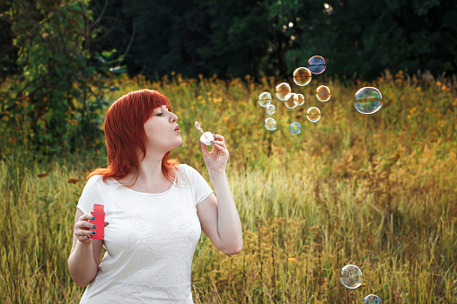 Young red-haired woman blowing soap bubbles. Happy girl in nature in the sun.