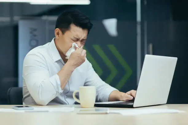 Photo of Sick man in office coughs and runny nose, Asian businessman with cold works in office on laptop