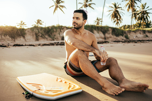 Young surfer sitting on sandy beach looking at the ocean with his bodyboard. Sport and water sport concept.