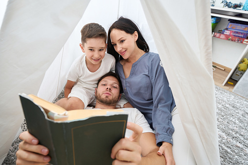 Dad, mom and son comfortably settled on the pillows, father is reading, mother and boy are listening enthusiastically
