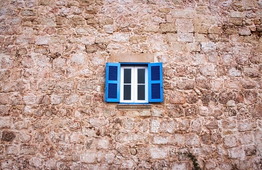 A window with white panes and blue wooden slats set in an old rustic wall