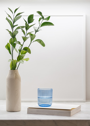Cropped view of colored glass with water, frame, plant in vase and book on table. Stylish kitchen tableware and flowers. Minimalism style. Home comfort. Objects composition idea. Cozy evening