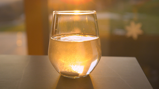 CLOSE UP: Effervescent tablet dissolving in sunlit glass of water. Sparkling beverage for regaining health backlit with beautiful golden light. Seasonal home treatment for immunity boost and health.